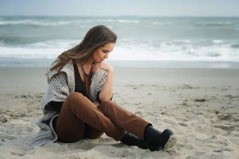 Eine traurige Frau in braunen Hosen und einem Pullover sitzt am Strand