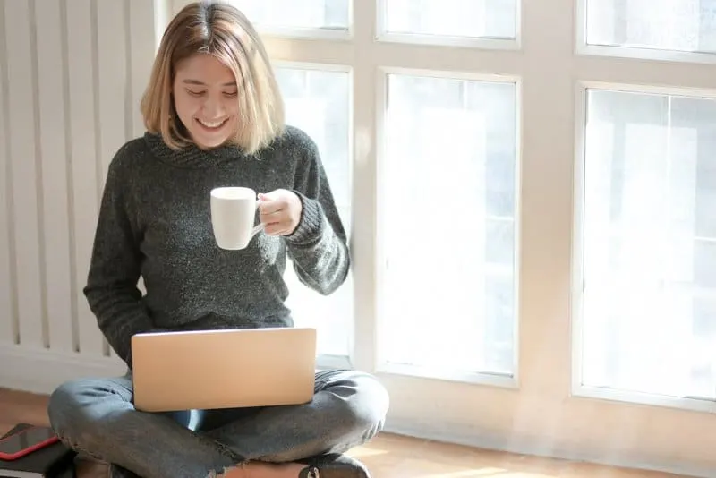 blonde Frau sitzt auf dem Boden, trinkt Kaffee und tippt auf Laptop