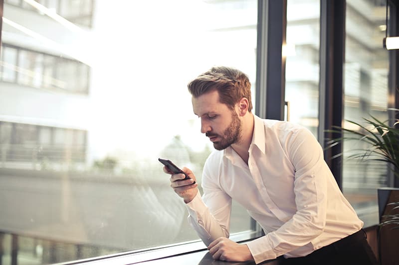 Ein Mann in einem weißen Hemd hält ein Smartphone in der Nähe des Fensters