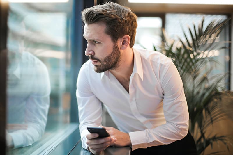 Ein ernster Mann hält sein Smartphone und schaut durch das Fenster