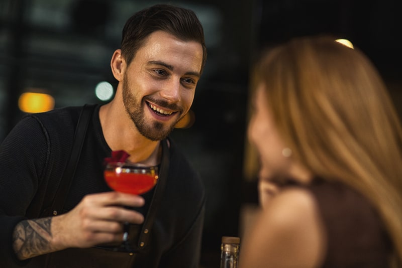 Ein hübscher Barkeeper lächelt, während er einer allein sitzenden Frau einen roten Cocktail anbietet