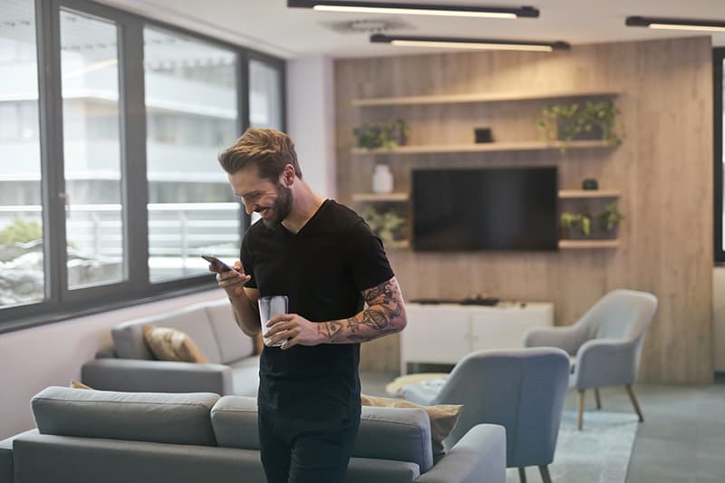 Ein lächelnder Mann benutzt sein Smartphone, während er im Wohnzimmer steht