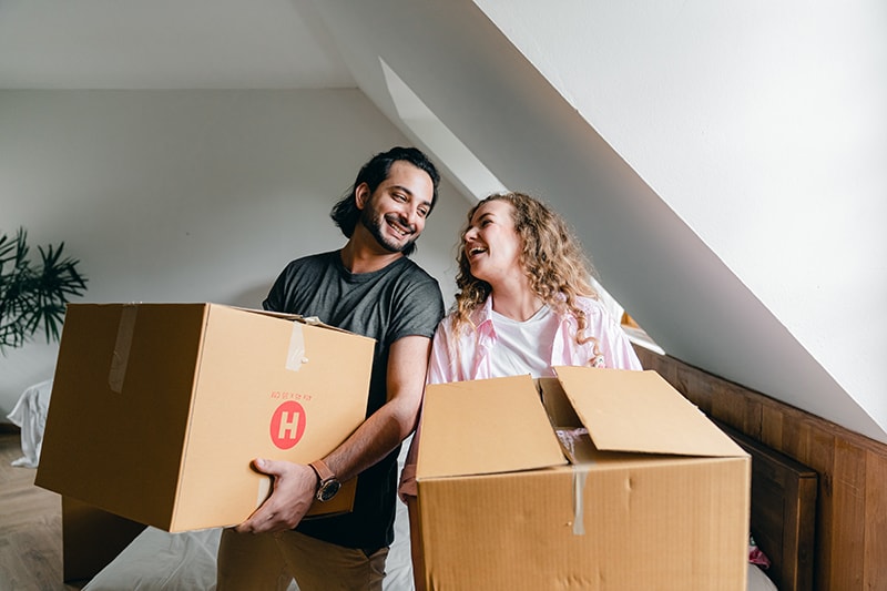 ein lächelndes Paar, das sich ansieht während Sie Kartons in der Wohnung halten