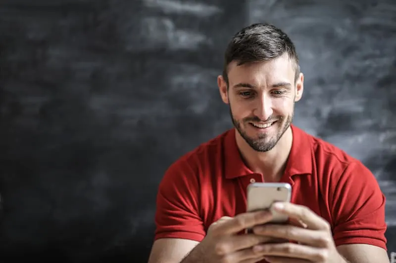 ein positiver Mann in einem roten Poloshirt mit einem Smartphone