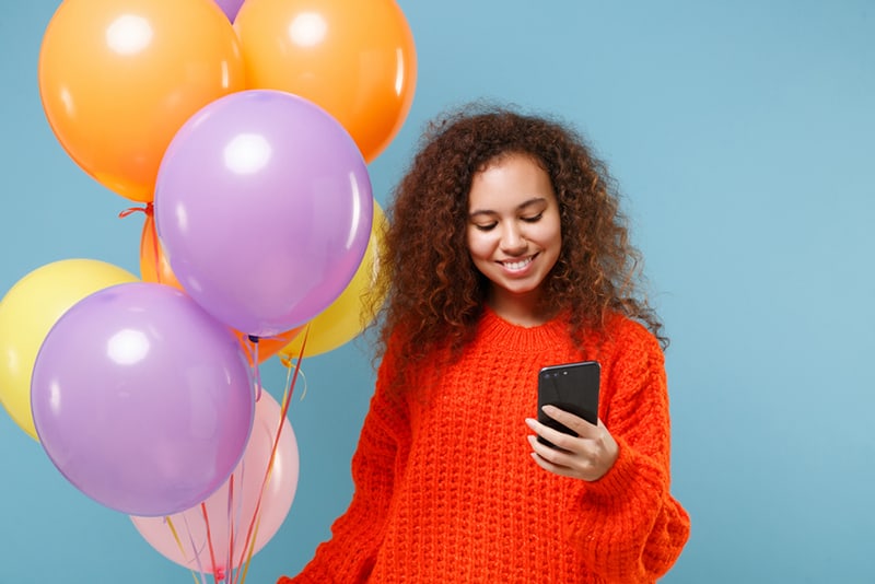 a woman celebrating a birthday holding a smartphone and standing near the balloons