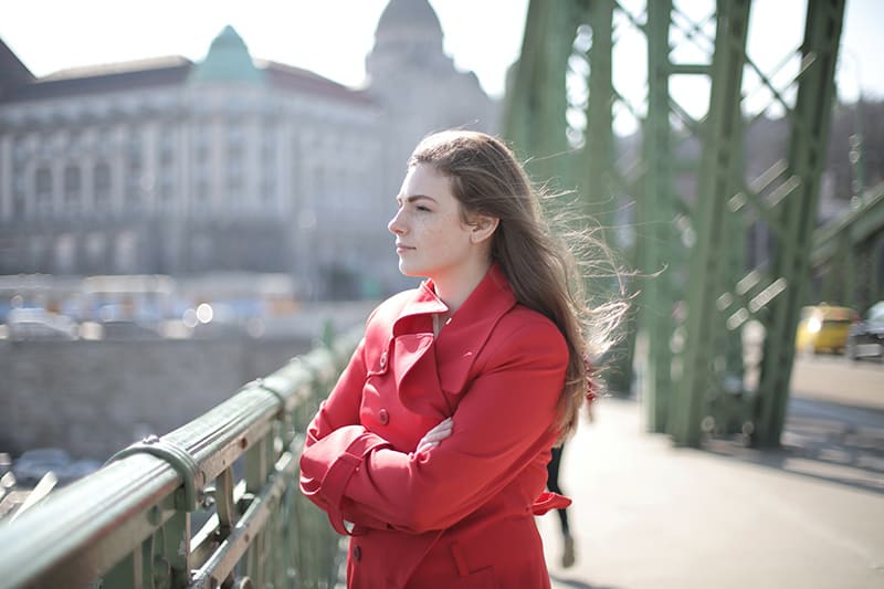 eine Frau in einem roten Mantel, die nahe Metallgeländern auf der Brücke steht und den Fluss betrachtet