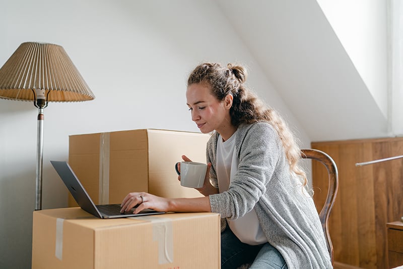 eine Frau, die einen Laptop auf dem Karton benutzt, während sie Sachen in der Wohnung organisiert