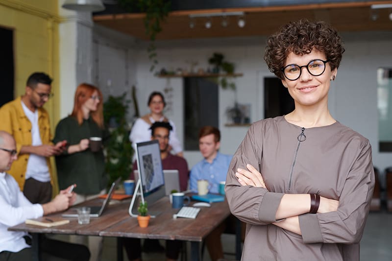 eine Geschäftsfrau, die mit verschränkten Armen im Büro steht, während ihre Kollegen hinter ihr arbeiten