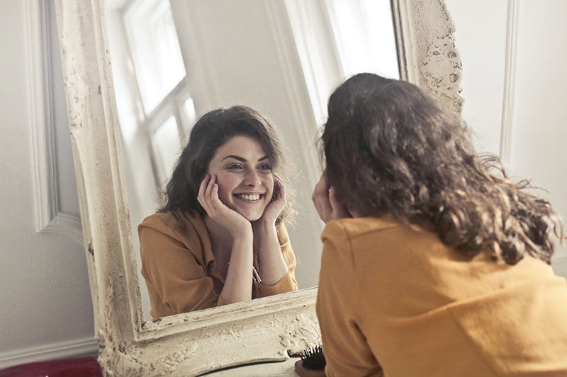eine lächelnde Frau, die sich in den Spiegel schaut, während sie sich auf das Regal lehnt