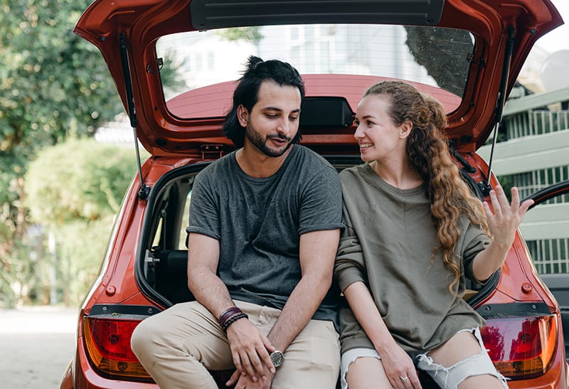 eine lächelnde Frau, die einem attraktiven Mann während eines Gesprächs beim Sitzen auf einem Autokofferraum zuhört