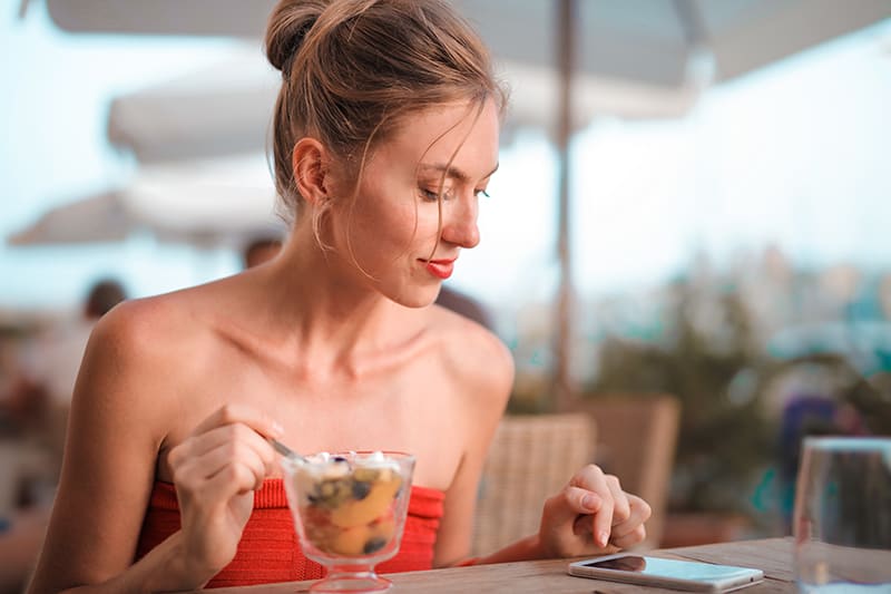 eine lächelnde Frau, die ihr Smartphone überprüft, während sie Obstsalat im Café isst