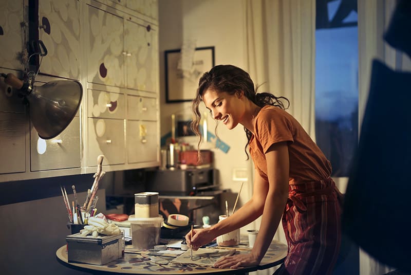 eine lächelnde Frau, die malt, während sie am Tisch im Atelier steht