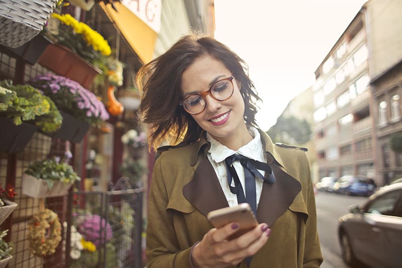 eine lächelnde Frau, die neben einem Blumenladen geht und ein Smartphone benutzt