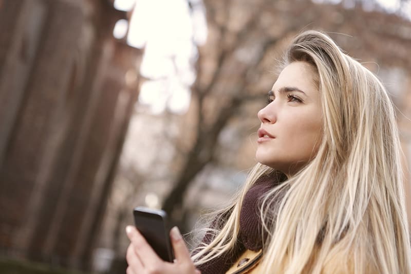 eine nachdenkliche Frau, die das Smartphone hält, während sie im Park steht und gerade schaut