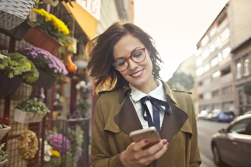 eine positive Frau, die ein Smartphone benutzt, während sie neben dem Blumenladen geht