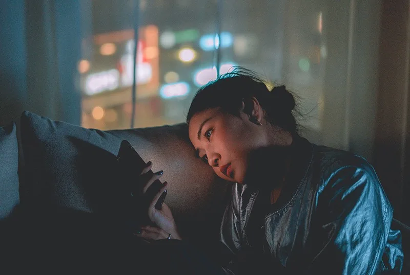 eine traurige Frau, die Smartphone betrachtet, während sie im Dunkeln auf der Couch sitzt