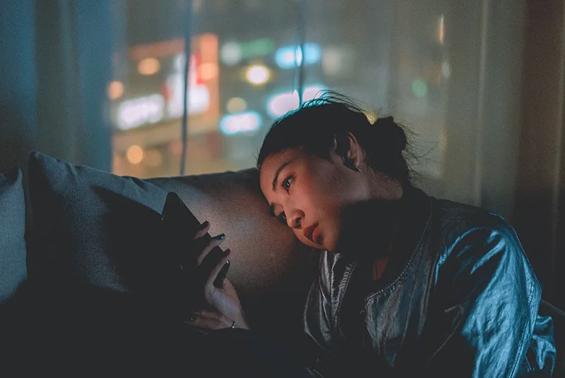 eine traurige Frau, die ein Smartphone betrachtet, während sie im Dunkeln sitzt