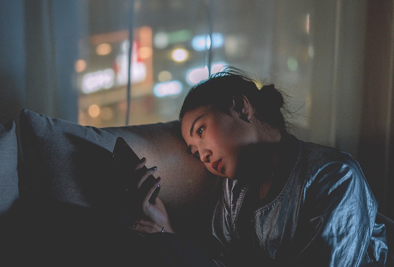 eine traurige Frau, die das Smartphone betrachtet, während sie im Dunkeln auf der Couch sitzt