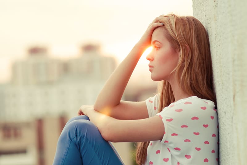 eine traurige Frau, die den Kopf berührt, während sie während des Sonnenuntergangs allein auf dem Dach sitzt