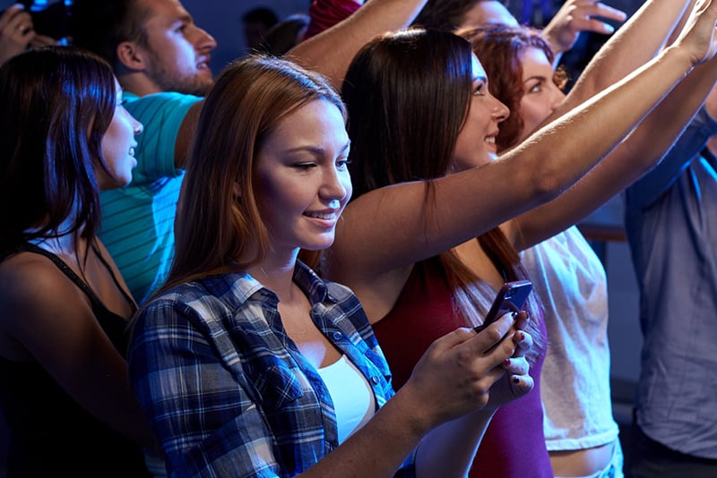 lächelnde Frau mit einem Smartphone beim Konzert im Club von Freunden umgeben