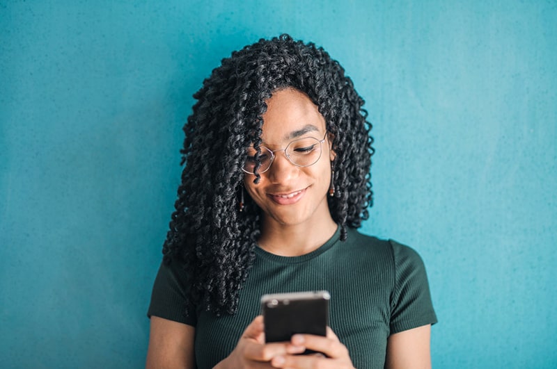 lächelnde Frau, die Brillen trägt und ein Smartphone benutzt, während sie nahe einer blauen Wand steht