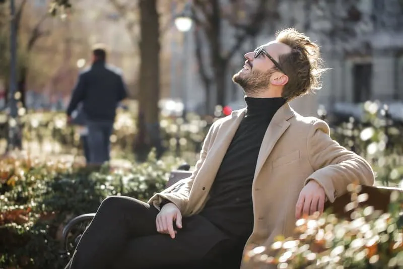 Auf einer Parkbank sitzt ein lächelnder Mann mit Brille und schaut zum Himmel hoch