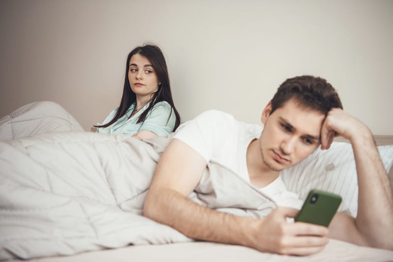 Eifersüchtige Frau, die seinen Freund mit einem Smartphone betrachtet, während sie zusammen auf dem Bett liegen