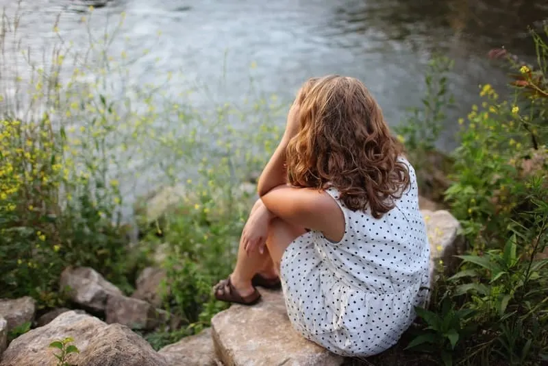 Ein Mädchen in einem weißen Tupfenkleid sitzt auf einem Felsen über dem Fluss