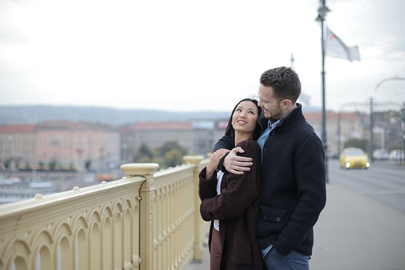 Ein Mann, der eine Frau von hinten umarmt und sich gegenseitig ansieht, während er auf der Brücke steht