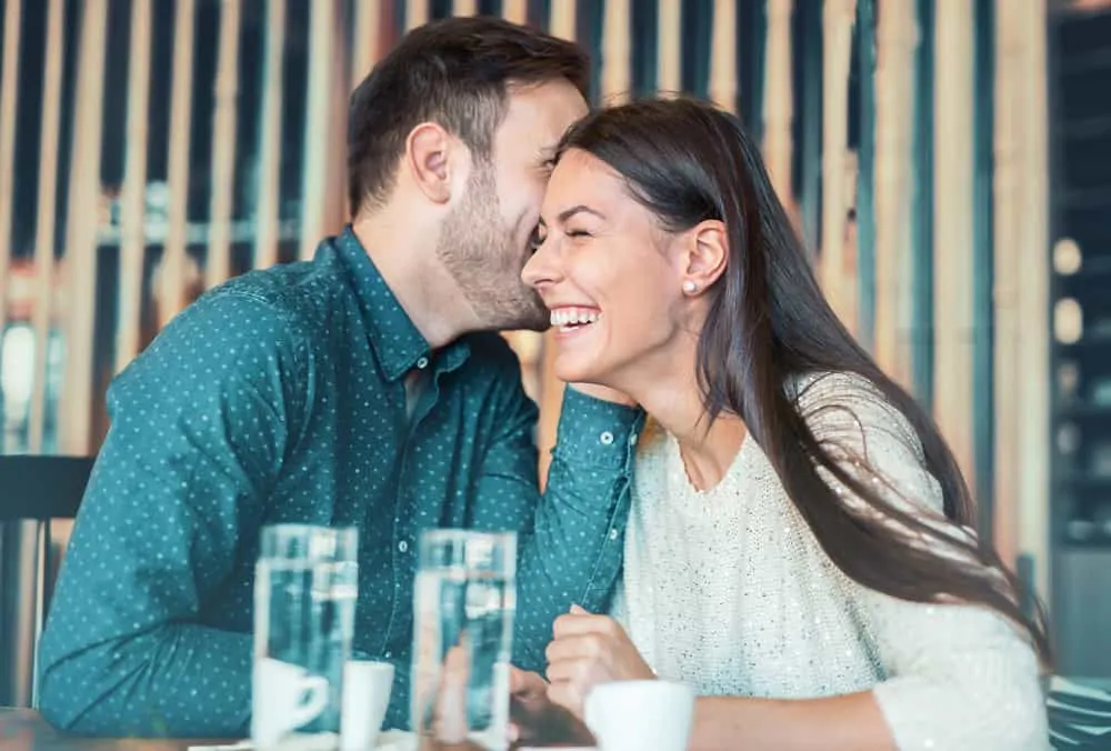 Ein Mann in einem Café flüstert einer lächelnden Frau etwas ins Ohr