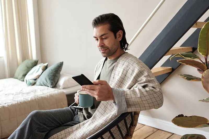 Ein Mann sitzt im Sessel im Schlafzimmer und schaut auf das Smartphone, während er Kaffee trinkt