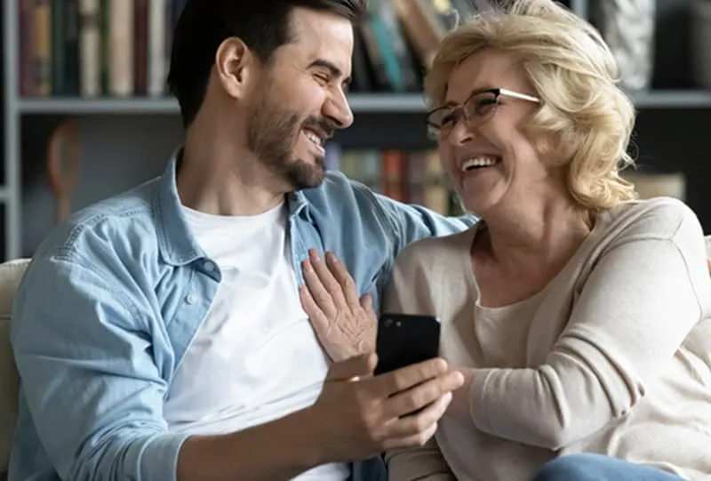 Ein Mann sitzt mit seiner Mutter auf der Couch und lacht, während er ein Selfie-Foto macht