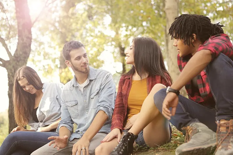 Ein Mann spricht und schaut zu einem lächelnden Mädchen, während er mit Freunden im Wald sitzt