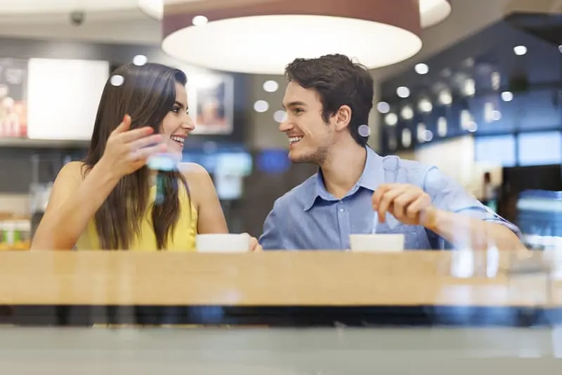 Ein lächelnder Mann hört einer Frau zu, die mit ihm spricht, während sie im Café Kaffee trinkt