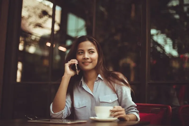 Eine lächelnde Frau in einem Hemd sitzt in einem Café und telefoniert