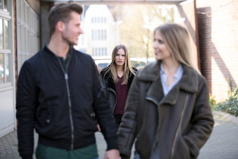 eine junge Frau, die eifersüchtig auf ein lächelndes Paar schaut, das vor ihr geht