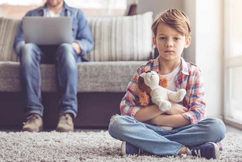 Trauriger kleiner Junge, der ein Spielzeug hält und auf dem Teppich sitzt, während sein Vater hinter ihm auf der Couch arbeitet