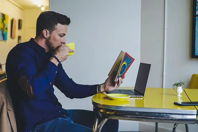 Ein Mann liest ein Buch und trinkt Kaffee, während er am Tisch sitzt