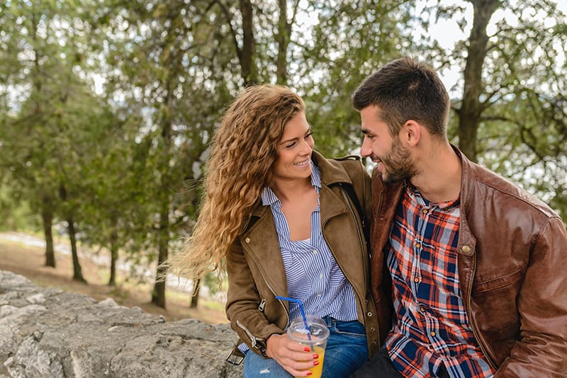 Ein Mann und eine Frau schauen sich an und lächeln, während sie auf der Betonwand im Park sitzen