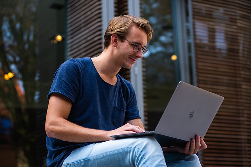 ein lächelnder Mann, der einen Laptop benutzt, während er vor dem Gebäude sitzt