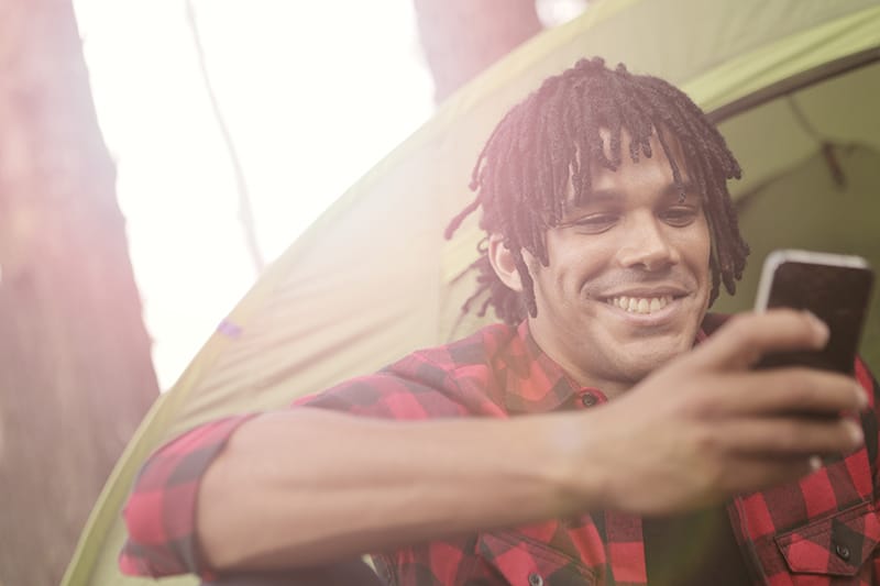 ein lächelnder Mann, der ein Smartphone benutzt, während er in der Nähe des Zeltes sitzt
