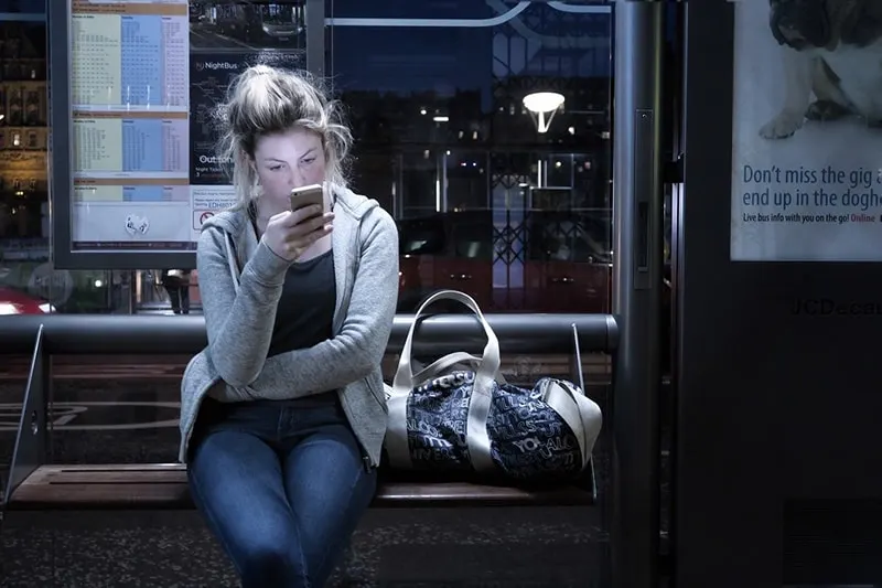 Eine Frau sitzt auf der Bank und versucht, einen Freund auf einem Smartphone an der Bushaltestelle zu kontaktieren