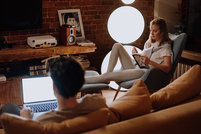 Eine Frau liest ein Buch und ein Mann benutzt abends einen Laptop im Wohnzimmer