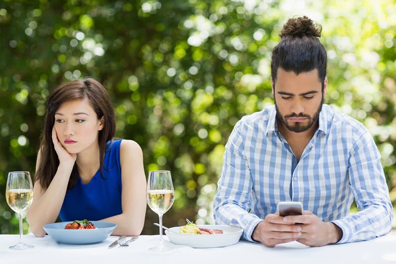 eine Frau, die gelangweilt aussieht, während sie in der Nähe eines Mannes sitzt, der während des Mittagessens im Restaurant ein Smartphone benutzt