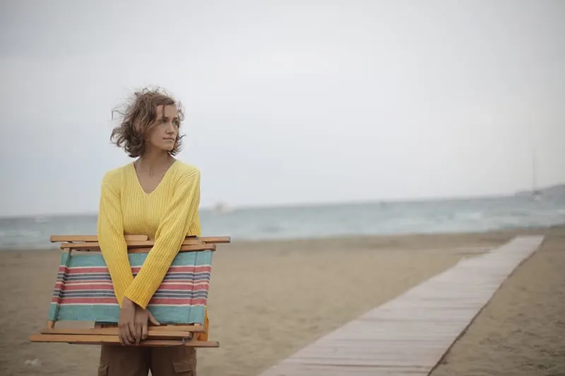 eine einsame Frau, die einen hölzernen Strandkorb trägt und nachdenklich aussieht, während sie auf dem Sand geht