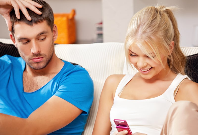 eine lächelnde Frau, die ein Handy benutzt, während sie in der Nähe ihres Freundes sitzt und auf ihr Handy schaut
