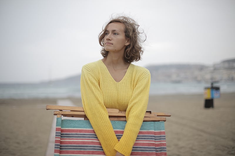 eine traurige Frau, die einen Liegestuhl hält, während sie allein am Strand steht