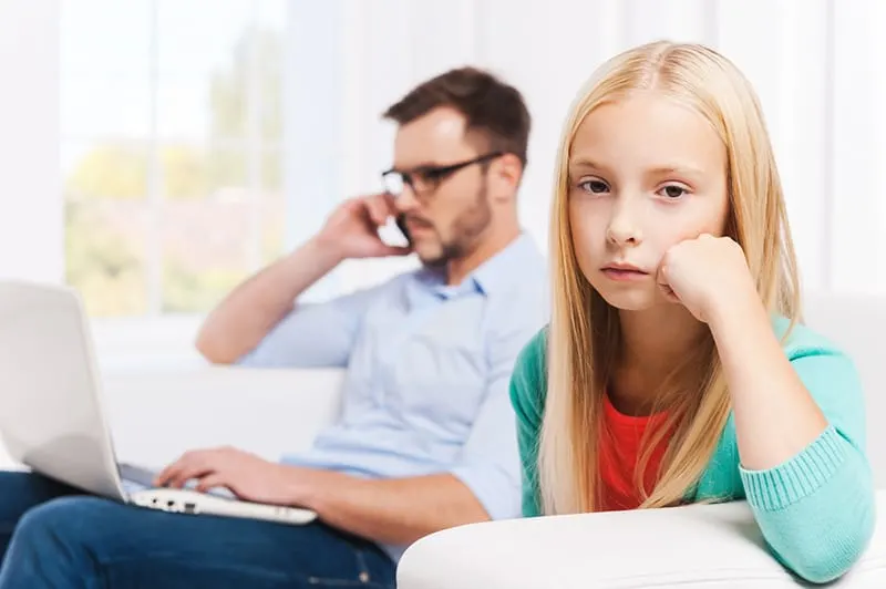 eine traurige kleine Tochter, die auf der Couch sitzt, während ihr Vater neben ihr am Laptop arbeitet