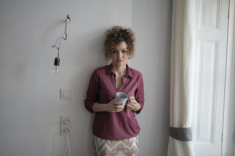 eine unzufriedene Frau, die eine Tasse Kaffee hält, während sie in der Nähe der Wand im Raum steht