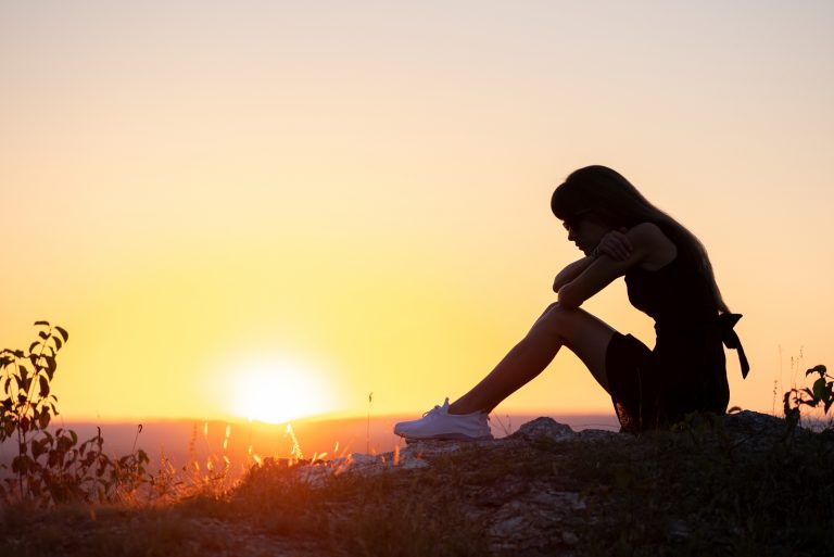 Auf dem Felsen bei Sonnenuntergang sitzt ein depressives Mädchen in einem schwarzen Kleid